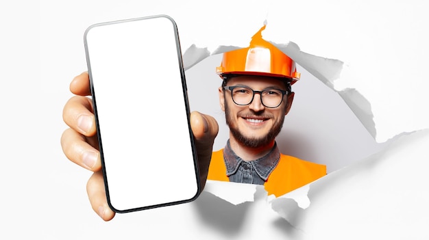 Studio portrait of young happiness man construction worker showing big smartphone with blank on screen through torn paper hole White background Wearing eyeglasses and safety equipment