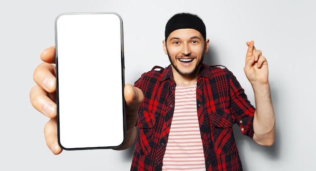 Studio portrait of young happiness man holding big smartphone with blank on screen in hand showing close to camera a device with mockup crosses finger on white background Wearing red shirt