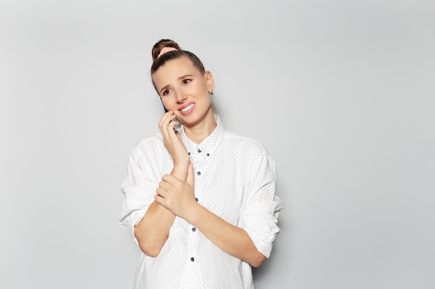 Studio portrait of young happy disappoint girl with hair bun talking on smartphone on grey background looking up wearing white shirt