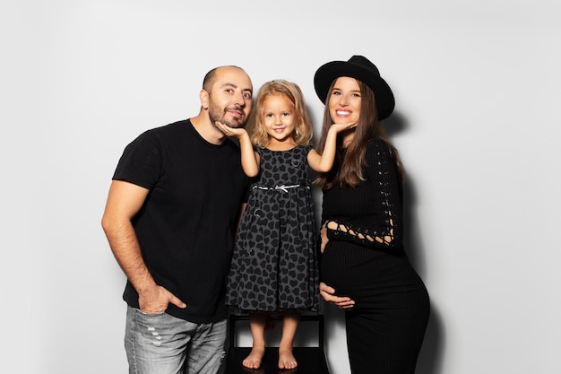 Studio portrait of young happy family daughter touching chins of her father and pregnant mother on white background Wearing hat dressed in black