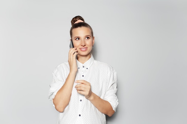 Studio portrait of young happy girl talking on smartphone on grey background looking up wearing white shirt