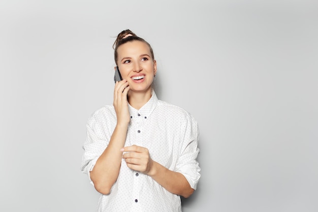 Studio portrait of young happy girl with hair bun talking on smartphone on light grey background looking up wearing white shirt