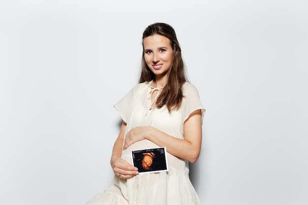 Studio portrait of young happy pregnant woman touching the belly showing ultrasound scan on white background