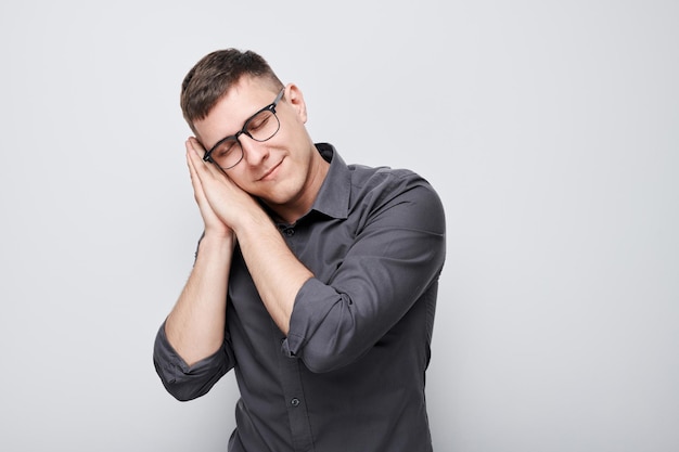 Studio portrait young man in glasses and shirt put palms under head pretending to sleep isolated on white background