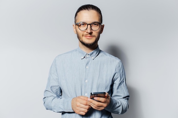 Studio portrait of young man holding smartphone in hands on light grey background Wearing eyeglasses and tshirt