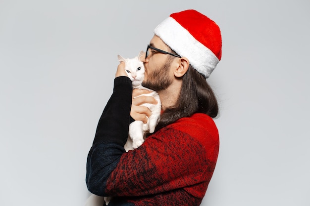 Studio portrait of young man in red Christmas sweater kisses and hugs a cat wearing Santa hat on white background