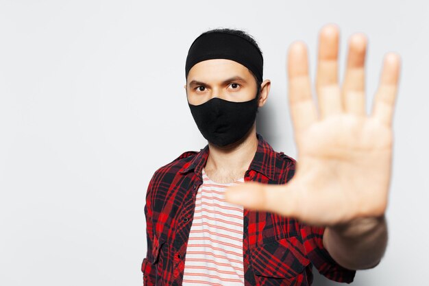 Studio portrait of young man wearing protective mask of black showing stop gesture by hand on white background