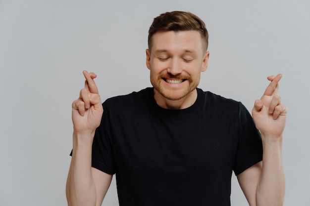 Studio portrait young man with closed eyes making wish, excited guy crossing fingers and asking for good luck while standing against grey studio background. Faith and superstition concept