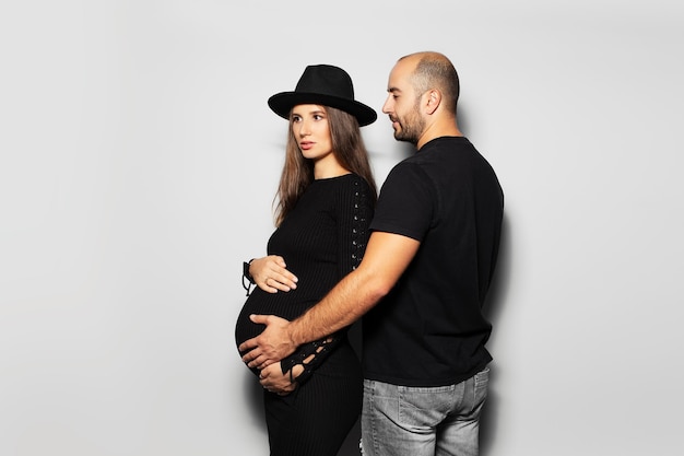 Studio portrait of young parents pregnant woman wearing black hat and dress man touching the belly on white background