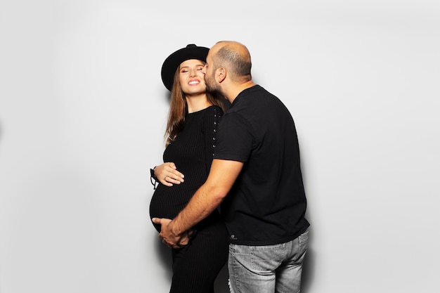 Studio portrait of young parents pregnant woman wearing black hat and dress man touching the belly on white background