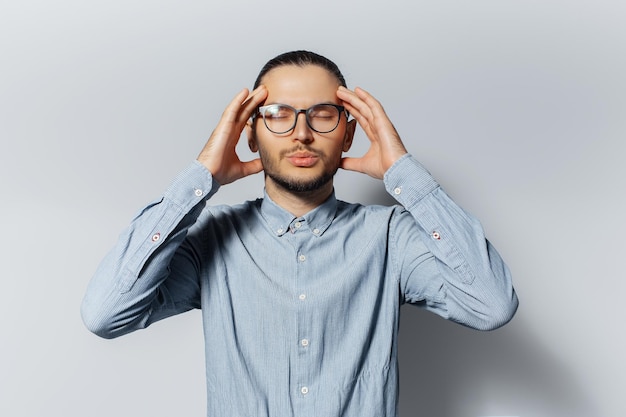 Studio portrait of young sad pensive man in blue shirt wearing eyeglasses touching his head with hands on white background