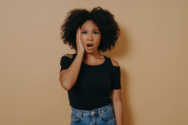 Studio portrait of young shocked african female with unexpected facial expression because of hearing bad news, mixed race woman keeping jaw dropped, wears casual outfit, posing against beige wall