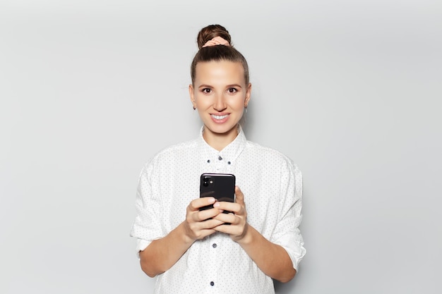 Studio portrait of young smiling girl using smartphone on grey background wearing white shirt