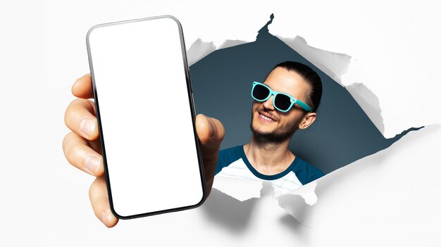 Studio portrait of young smiling guy showing big smartphone with blank on screen through torn paper hole White and blue background Wearing cyan sunglasses