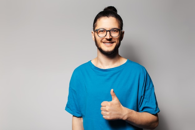 Studio portrait of young smiling man in blue shirt showing thumb up on grey