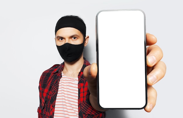 Studio portrait of young smiling man holding big smartphone with blank on screen in hand showing close to camera a device with mockup wearing medical protective mask of black on white background