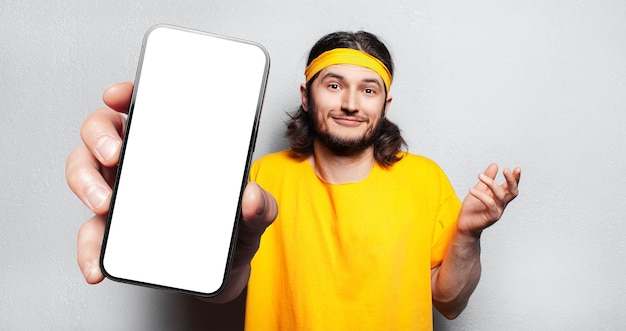 Studio portrait of young smiling man holding big smartphone with blank on screen in hand showing close to camera a device with mockup on white background Wearing yellow shirt and band on head