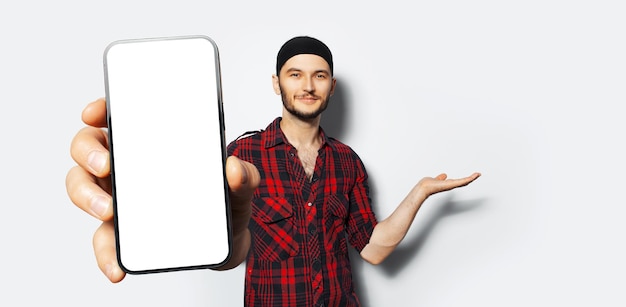 Studio portrait of young smiling man holding big smartphone with blank on screen in hand showing close to camera a gadget with mockup point hand presenting or demonstrate with palm on white