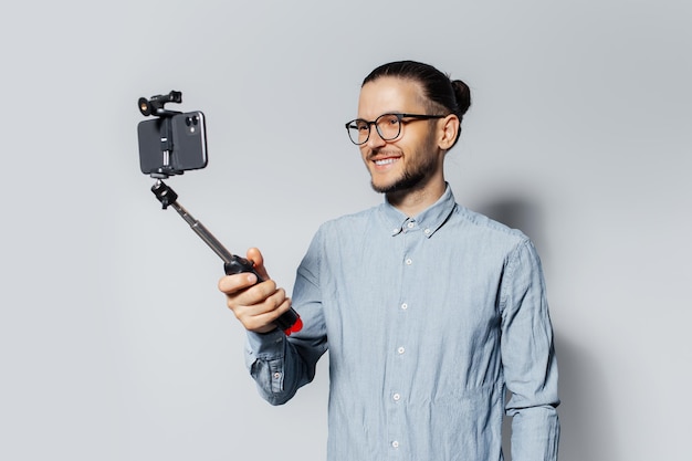 Studio portrait of young smiling man taking selfie photo via smartphone and stick on white background Wearing eyeglasses and blue tshirt