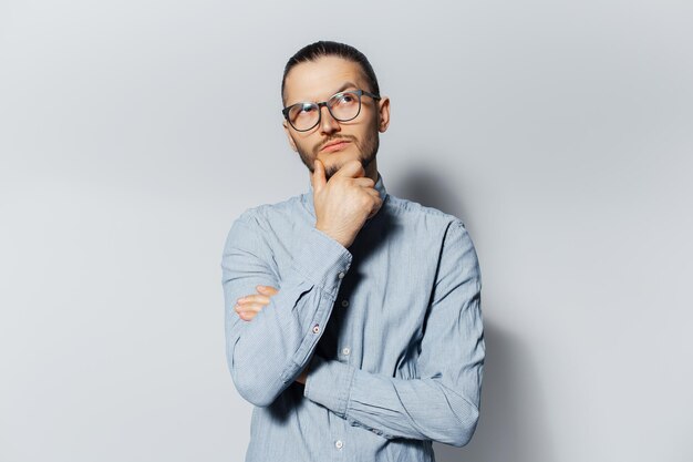 Studio portrait of young thoughtful man touches chin looking up on white background Wearing eyeglasses and blue shirt New idea concept