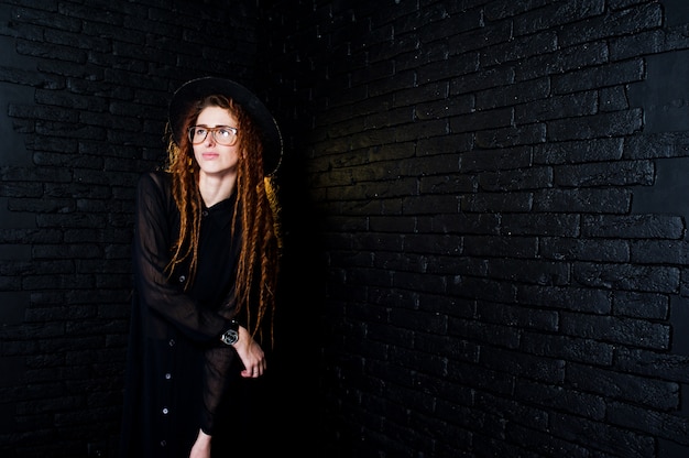 Studio shoot of girl in black with dreads, at glasses and hat on brick background.