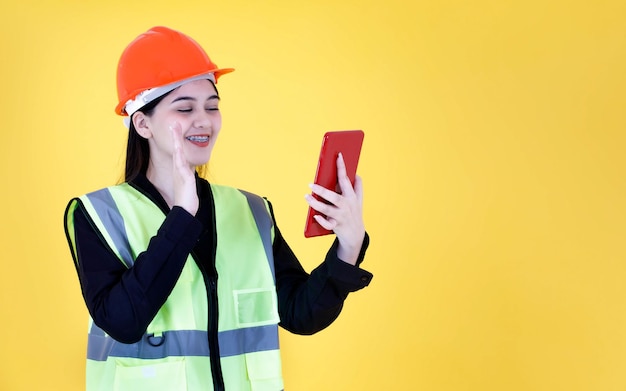 Studio shot Asian female professional engineering foreman manager with braces in hard helmet and reflective safety vest smiling greeting say hi hello goodbye on tablet video call on yellow background.