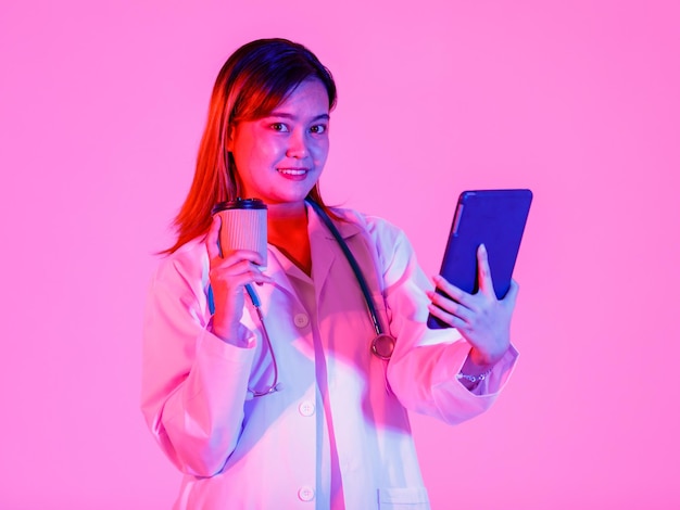 Studio shot of Asian successful professional confident female doctor in lab coat uniform hanging stethoscope around neck take coffee break standing smiling holding tablet computer on pink background.