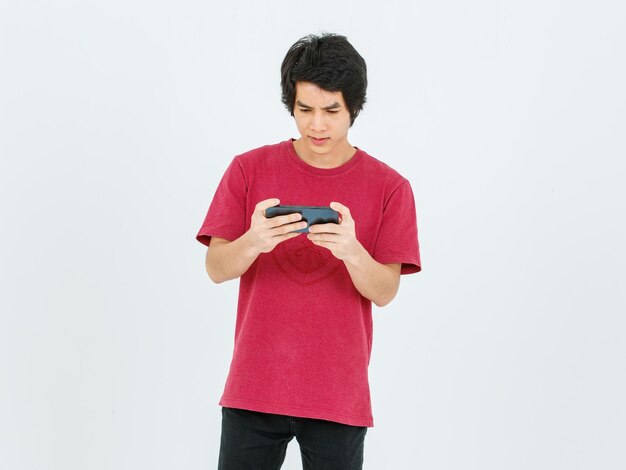 Studio shot of Asian young moody confused stressed teenager male model in casual street style outfit standing holding touchscreen smartphone in hands playing streaming game online on white background