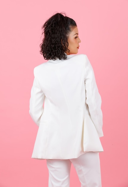 Studio shot of Asian young sexy curly hairstyle professional successful businesswoman in white fashionable casual suit standing posing holding hands in pants pockets turning back on pink background