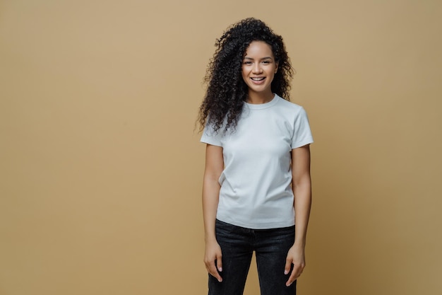 Studio shot of attractive cheerful woman with Afro hairstyle smiles positively rejoices buying new clothes wears white t shirt and jeans enjoys free time for rest poses against brown background