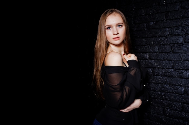 Studio shot of brunette girl in black blouse with bra and shorts against black brick wall.