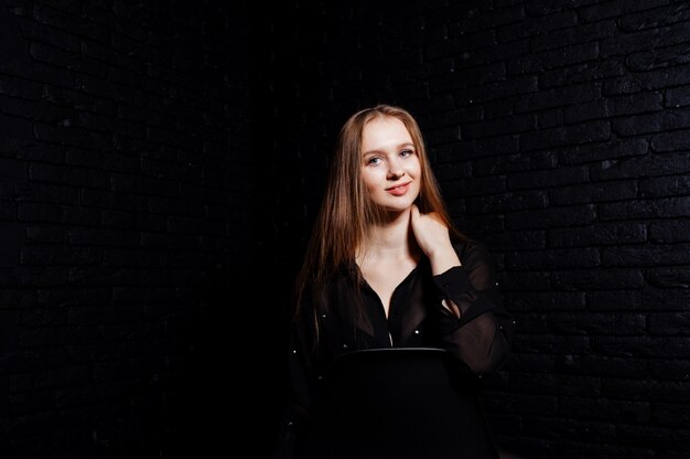 Studio shot of brunette girl in black blouse with bra and shorts against black brick wall.