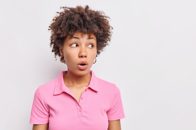 Studio shot of curly haired young woman looks away mysteriously holds breath keeps mouth opened dressed in casual pink t shirt isolated over white wall