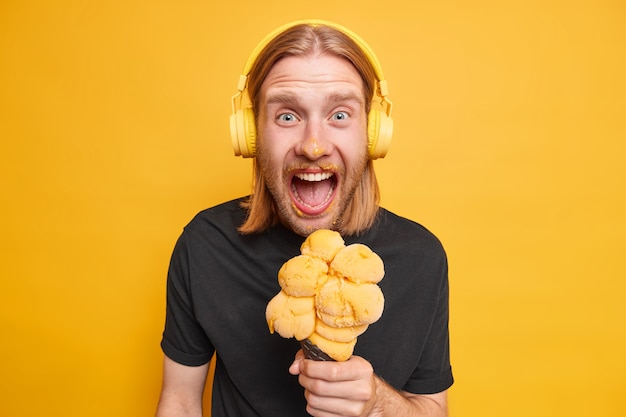 Studio shot of emotional redhead guy dirty after eating cone ice cream enjoys yummy summer dessert effords himself tasty food listens audio track in headphones