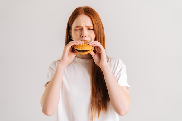 Studio shot of happy hungry young woman with closed eyes enjoying bite of appetizing delicious hamburger on white isolated background Closeup front view of female eating tasty burger