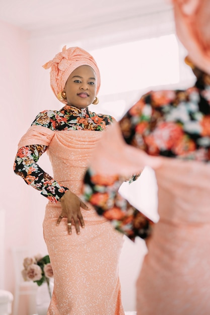 Studio shot of joyful pretty African lady wearing stylish pink dress with traditional print and teadwrap, posing near big mirror looking at reflection with smile.