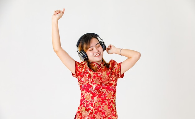 Studio shot of millennial Asian female model in red Chinese traditional cheongsam qipao shirt closed eyes standing listening to music via headphone headset dancing raised hands up on white background.