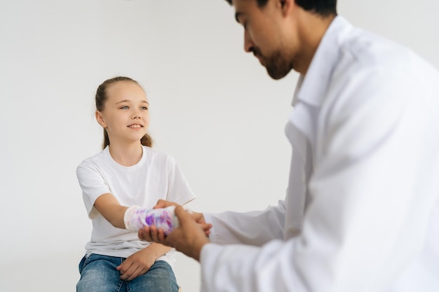 Studio shot of positive pediatrician male doctor in uniform consulting smiling little girl patient with broken hand wrapped in white plaster bandage