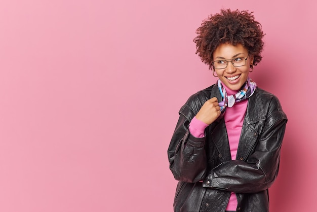 Studio shot of pretty curly haired young woman smiles happily looks away wears black leather jacket and kerchief around neck has glad expression isolated over pink background blank copy space