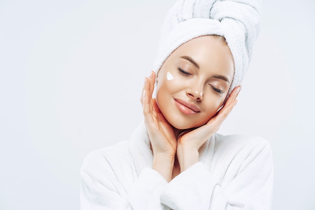 Studio shot of relaxed beautiful woman with closed eyes tilts head, touches skin gently, applies face cream, wears bath towel on head after taking spa procedures, enjoys facial treatments, body care