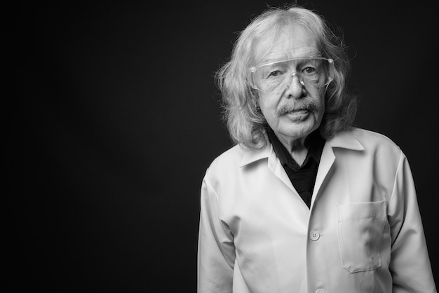 Studio shot of senior man doctor with mustache wearing protective glasses against gray wall