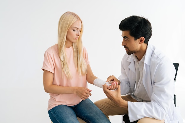 Studio shot of serious male practitioner orthopedic in uniform examining broken hand wrapped in plaster bandage of injured blonde female patient at checkup