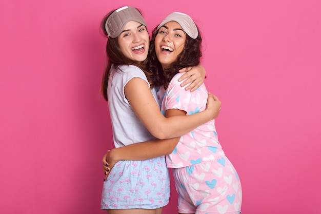 Studio shot of smiling adorable women with sleep masks, wearing pajamas, stands hagging isolated over rose studio background, ladies hagging each other, expressing happyness. Friendship concept.