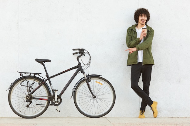 Photo studio shot of young male with curly hair, dressed in fashionable anorak, points with index finger at bicycle, advertises new model, drinks takeaway cooffe, makes choice, isolated on white background.