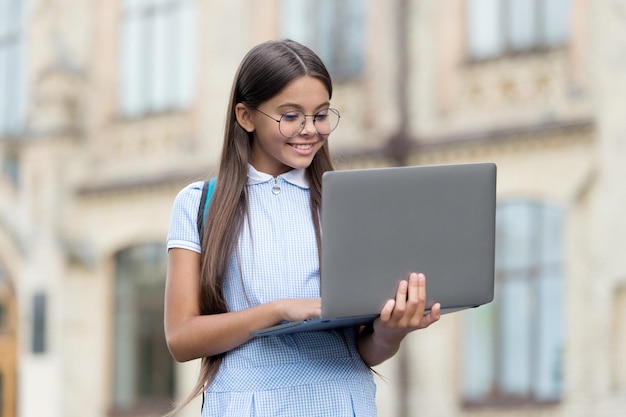 Study cheerful kid working on laptop online education back to school teen girl use computer in school yard child with backpack and notebook new technology in modern life Purchasing