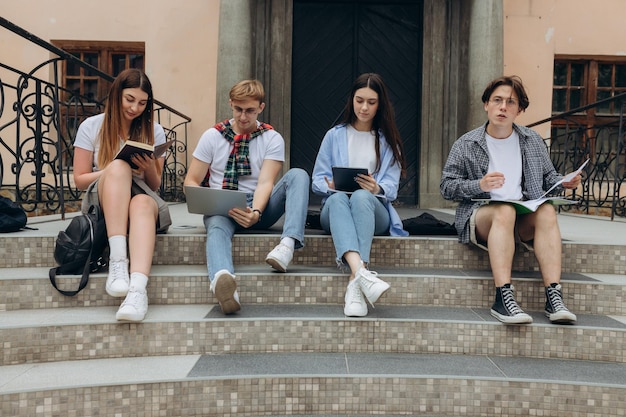 Study together is fun teamwork team building concept Four happy students are sitting near college building and holding books devices wearing casual smart smiling on a nice summer