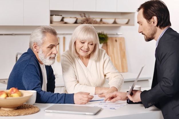 Studying details. Happy concentrated old couple sitting at home and discussing contract with real estate agent while exchanging opinions