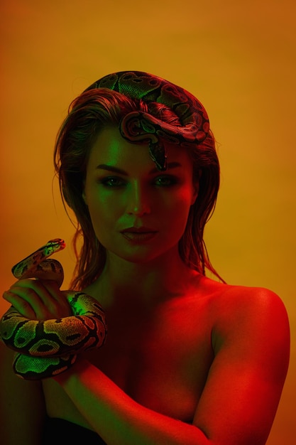 Stunning woman in a mix of red and green light with python snake as a crown atop her head and gracefully holds another in her hand