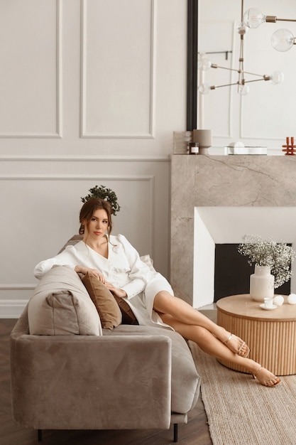 A stunning young brownhaired woman in a white cocktail dress sits on the sofa in the classic white i...