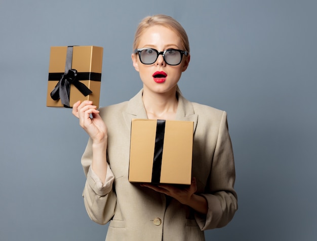 Style blonde in jacket and glasses holds gift box on gray space